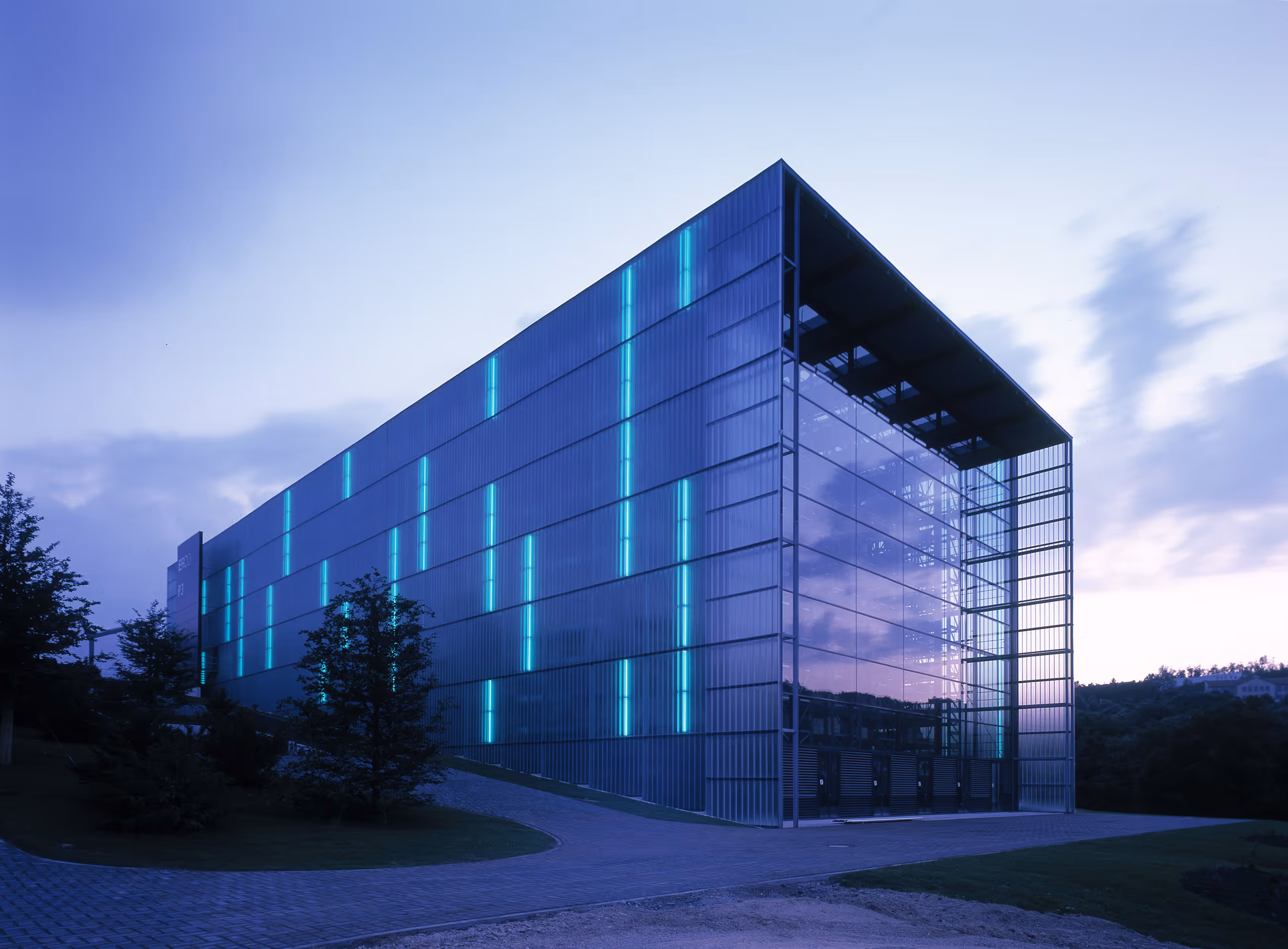 Modernes Gebäude mit reflektierenden Glaswänden und vertikalen blauen Lichtern bei Dämmerung.