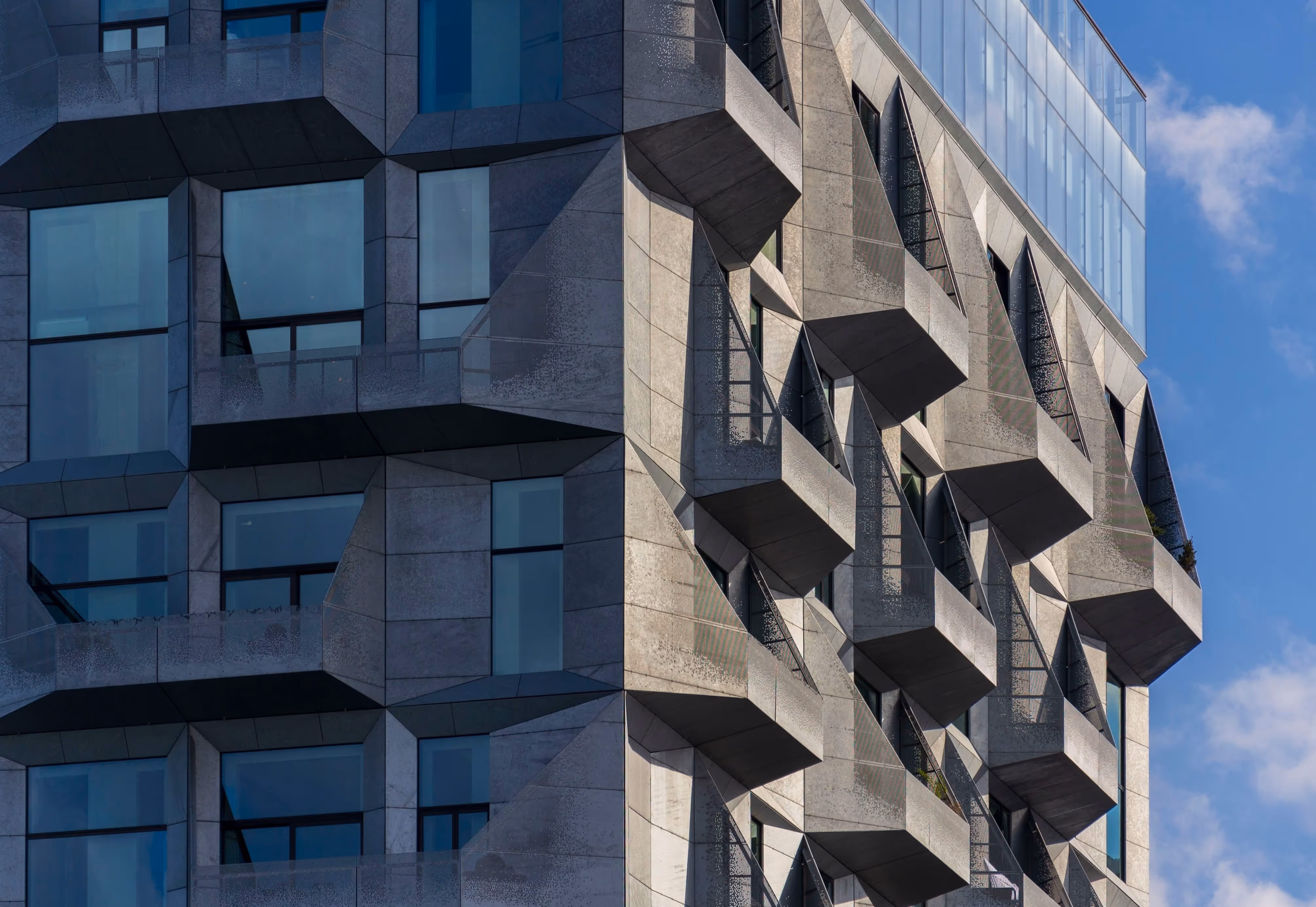 Moderne Fassade mit geometrisch geformten Balkonen aus Beton und großen Glasfenstern unter blauem Himmel.