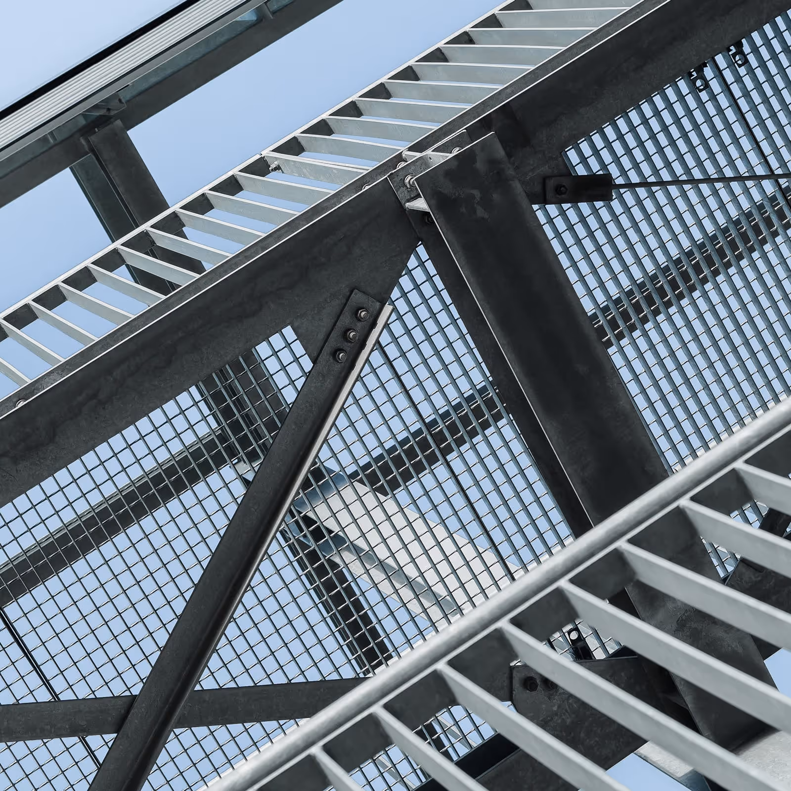 Ansicht von unten auf ein verzinktes Stahlgitter und Treppengeländer vor blauem Himmel.