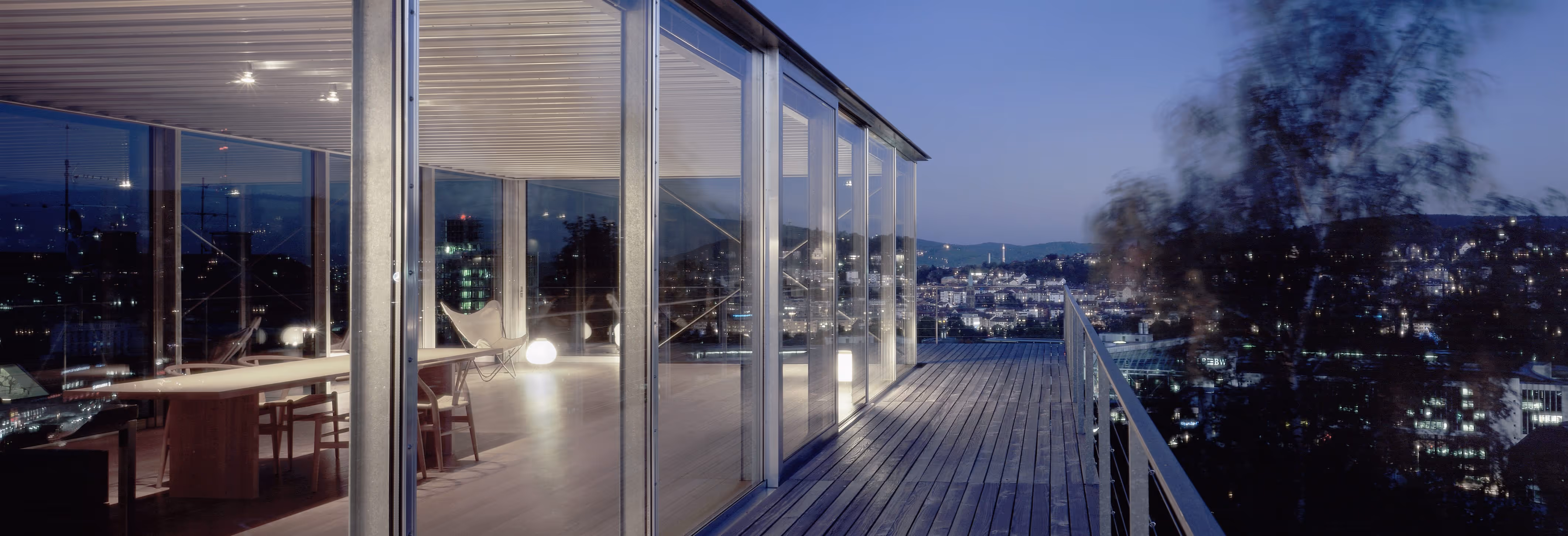 Abendaufnahme eines modernen Glaspavillons mit Holzboden und städtischer Beleuchtung im Hintergrund.