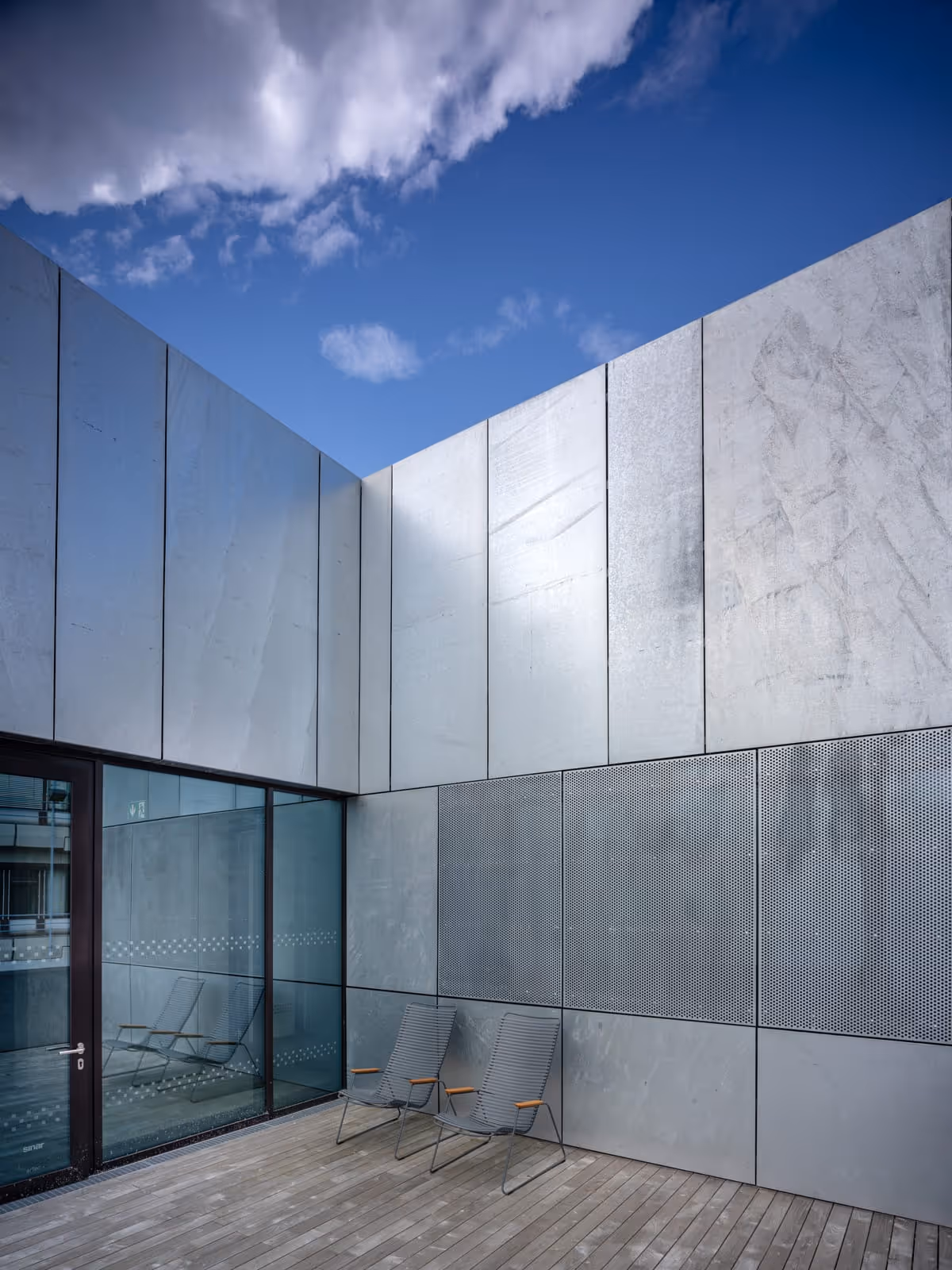 Zwei graue Stühle auf einer Holzterrasse vor einer modernen Fassade aus grauen Metallplatten und Fenster mit Reflexion.