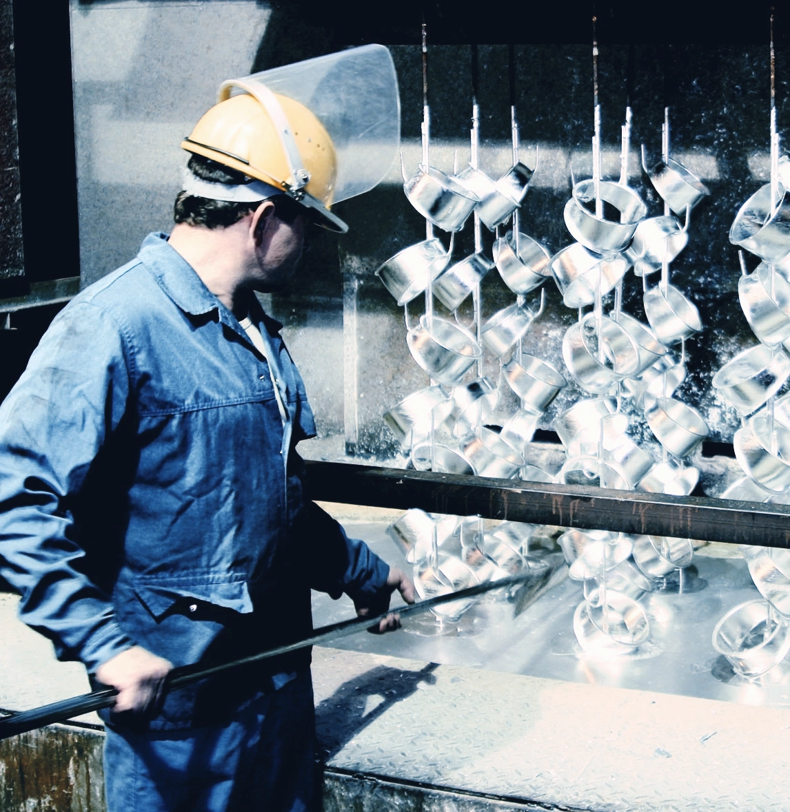 Arbeiter mit Schutzhelm und Gesichtsschutz überwacht das Eintauchen glänzender Metallteile in ein Feuerverzinkungsbad.