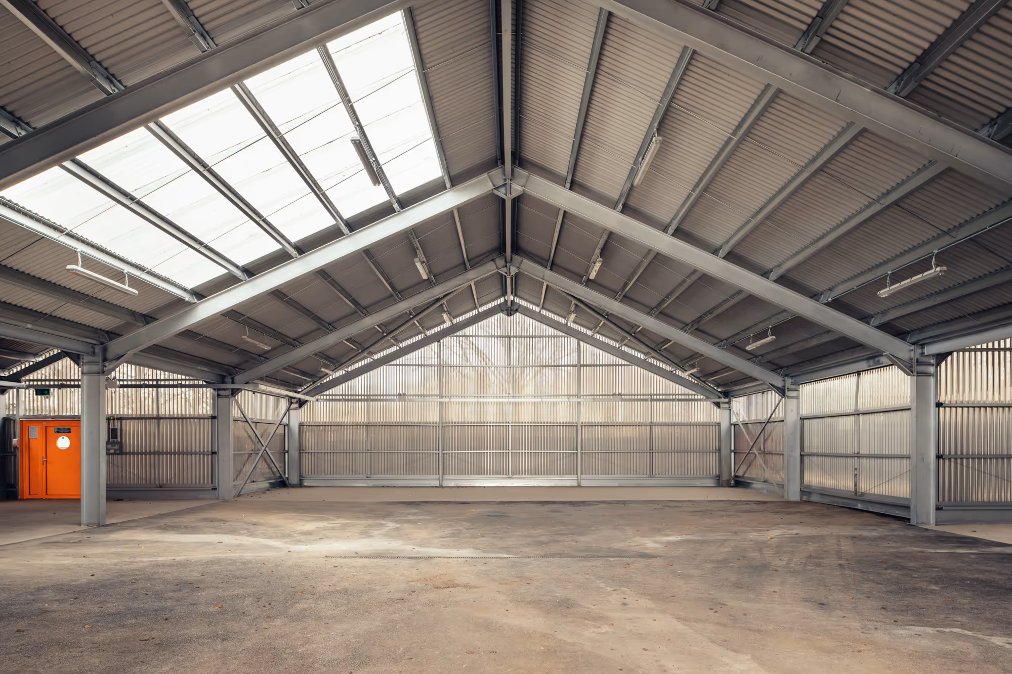 Leerer, großer Industriehallenraum mit Metallstruktur, grauen Wänden und einer orangefarbenen Tür auf der linken Seite.