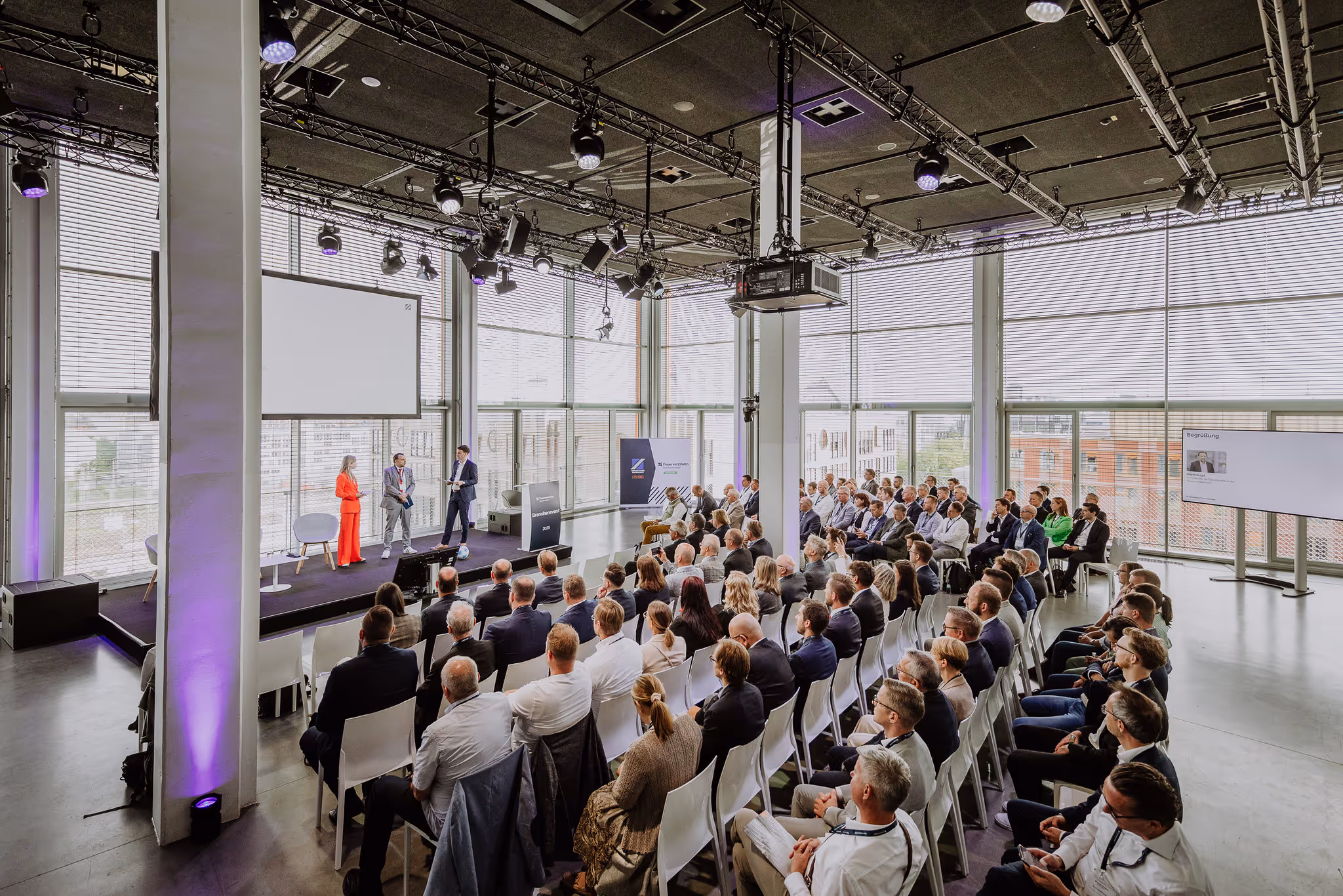 Geschäftskonferenz mit einem vollen Publikum, drei Personen präsentieren auf einer Bühne in einem modernen Veranstaltungsraum mit großen Fenstern.