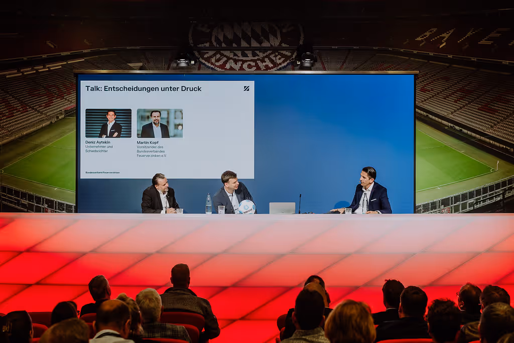 Drei Männer sitzen an einem Tisch auf einer Bühne vor einem Bildschirm mit dem Titel 'Talk: Entscheidungen unter Druck' vor einem Publikum in einer Sportarena.