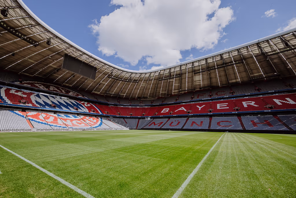 Das leere Fußballstadion Allianz Arena mit grünem Spielfeld und sitzenden Bereichen, die das Logo und den Namen von FC Bayern München zeigen.
