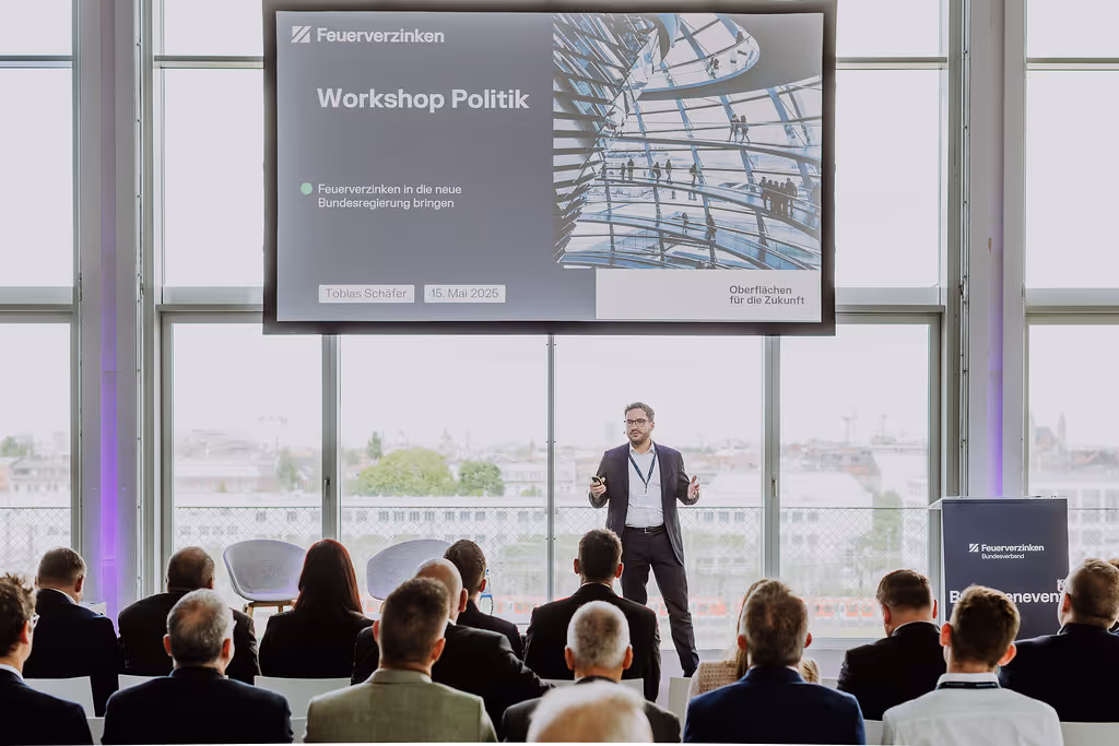 Ein Mann hält einen Vortrag vor einem Publikum in einem hellen Raum mit großen Fenstern, auf der Leinwand steht „Workshop Politik“.