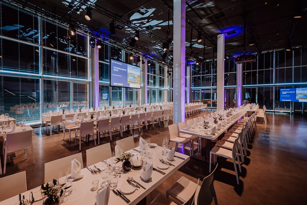 Ein großer Veranstaltungsraum mit langen, elegant gedeckten Tischen und Stühlen, beleuchtet mit violetten Akzenten und großen Fenstern im Hintergrund.