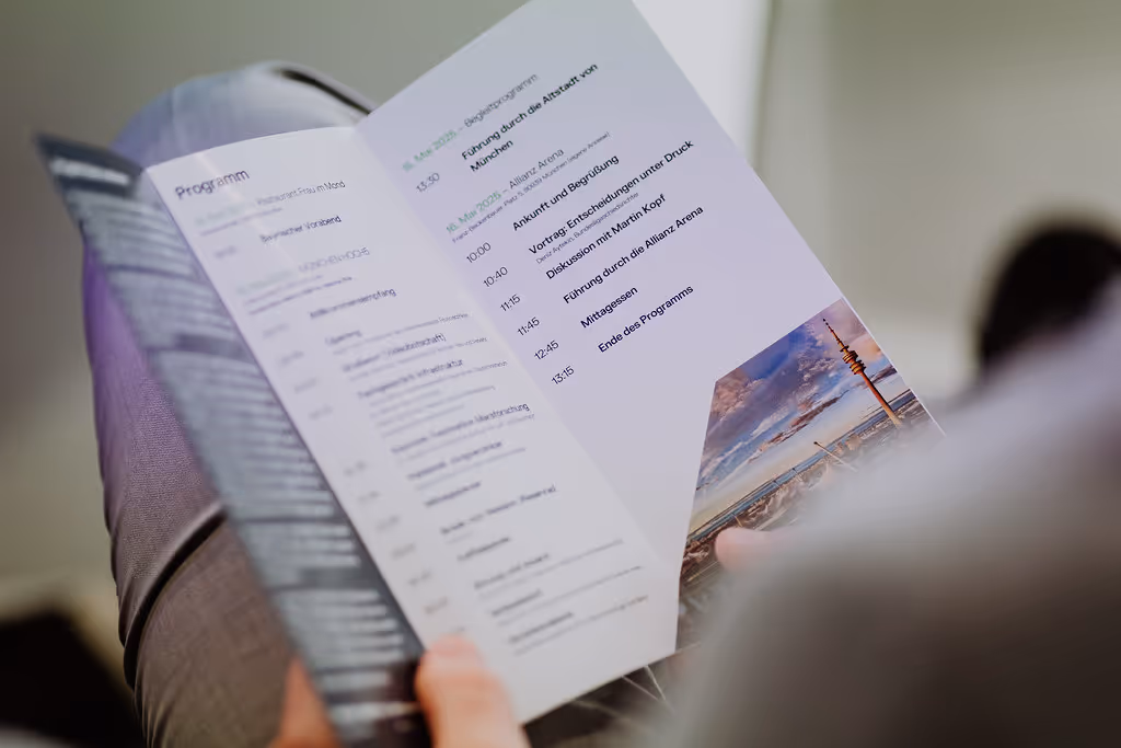 Person hält einen Programmflyer mit Zeitplan und Veranstaltungsdetails sowie einem Bild des Münchner Fernsehturms.