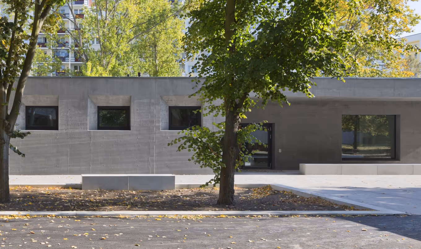 Moderner niedriger Betonbau mit Fensterfront, umgeben von Bäumen mit herbstlichen Blättern.