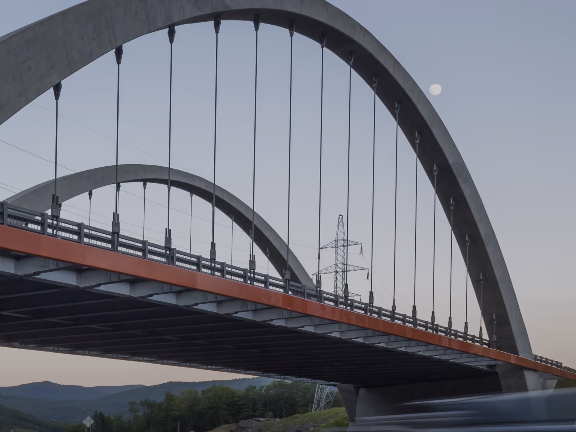 Bogenbrücke mit Stahlseilen bei Dämmerung, im Hintergrund Hügel und Vollmond.