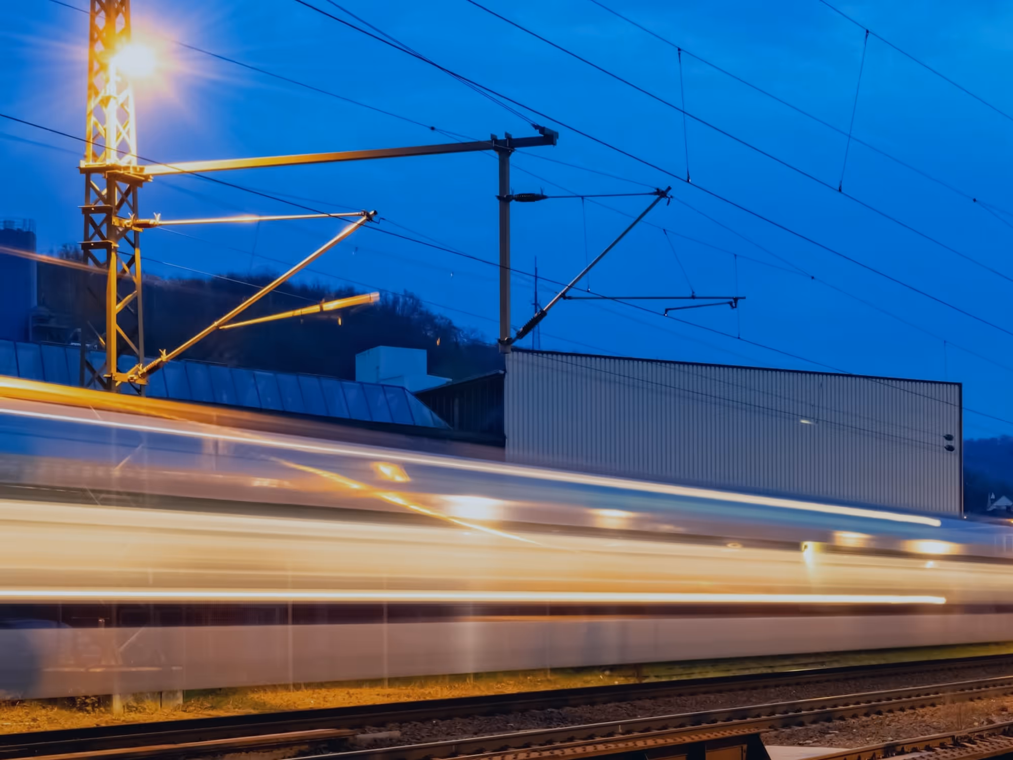 Ein schnell fahrender Zug mit Lichtspuren auf einem Gleis bei Abenddämmerung vor einem Industriegebäude.