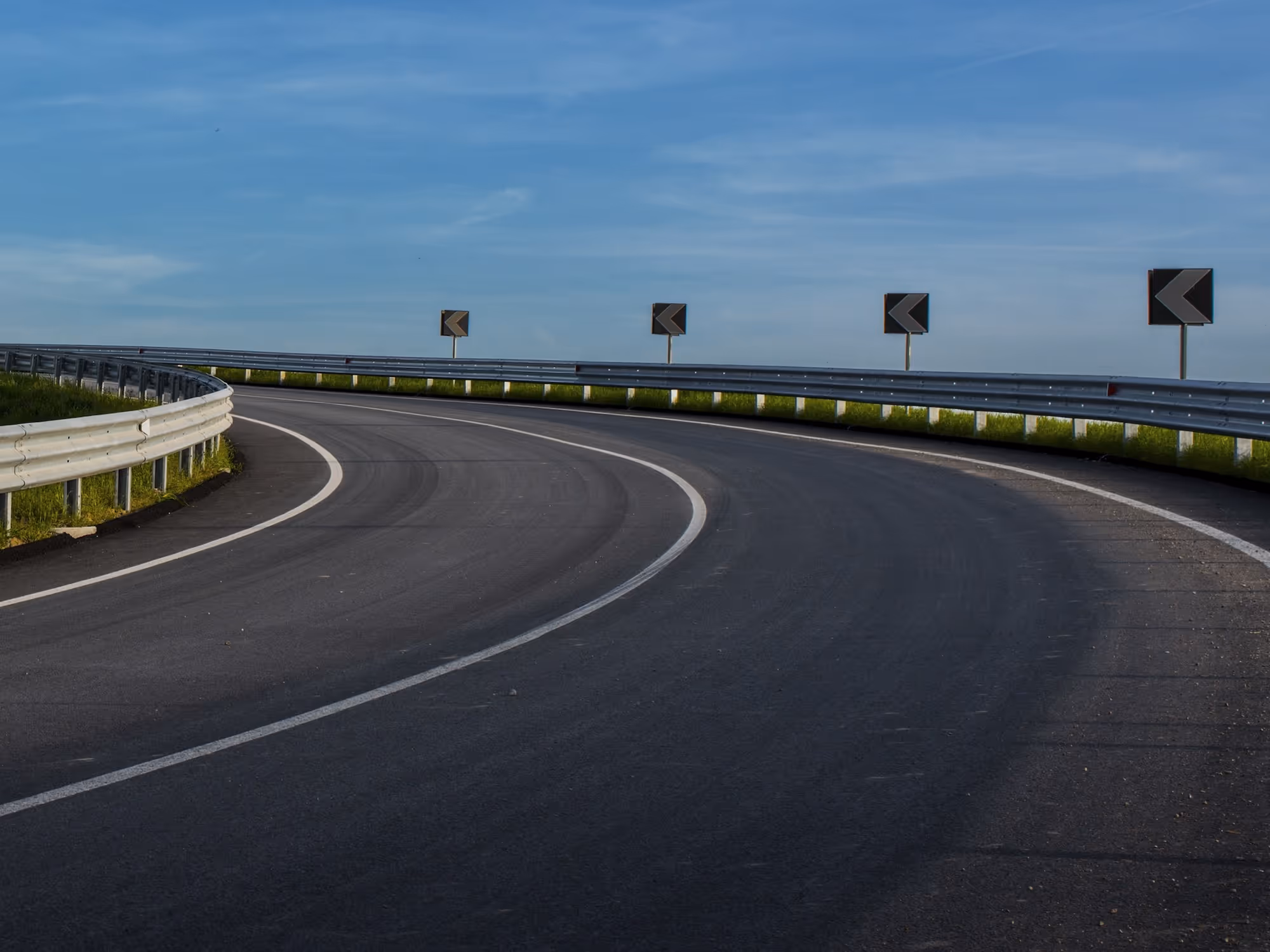 Kurvige Asphaltstraße mit Leitplanken und Linkspfeilschildern vor blauem Himmel.
