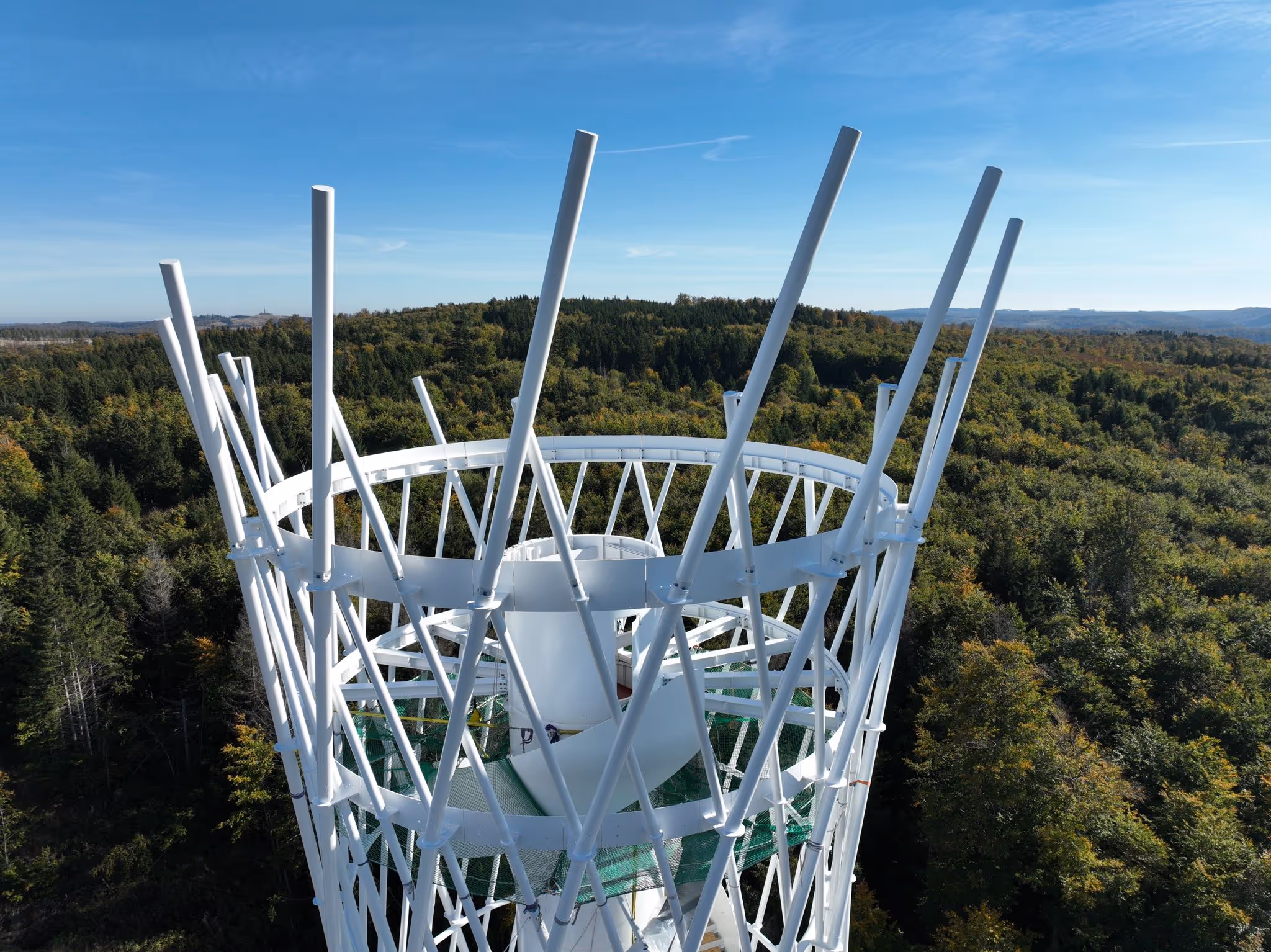 Blick von einer weißen, modernen Aussichtsplattform mit Stahlkonstruktion auf einen dichten grünen Wald unter klarem blauem Himmel.