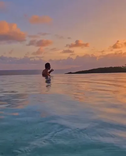 A person in an infinity pool at sunset, the sky reflecting orange, pink, and purple hues, with the sea and coastline in the distance. A luxury scene overlooking the sea in Guadeloupe.
