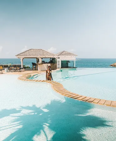 Woman in swimsuit in an infinity pool with a curved wooden path, facing beachfront bars and the ocean. Represents a 5 star luxury hotel in Guadeloupe.