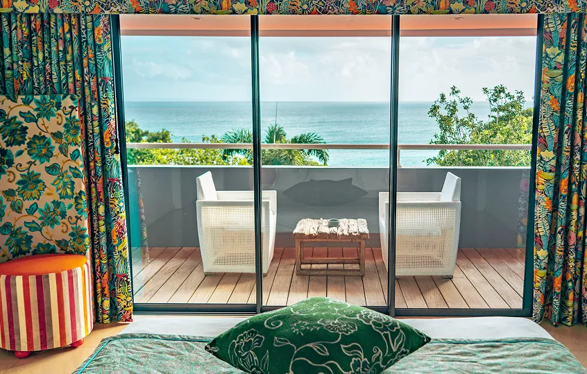 Luxury hotel room with a comfortable bed and a green cushion, featuring large windows opening onto a balcony with chairs, offering panoramic views of the Caribbean Sea and tropical vegetation. Ideal for a stay overlooking the sea in Guadeloupe.