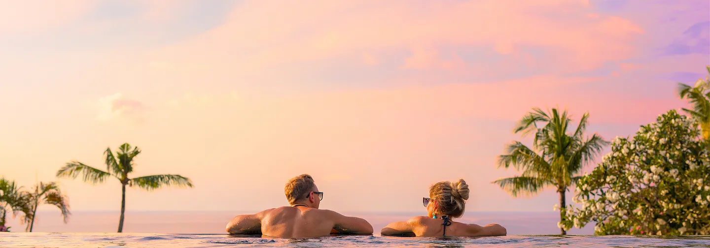 A couple relaxes in an infinity pool at sunset, taking in the pink and orange sky and the sea in the distance, with palm trees in silhouette. This scene evokes a romantic stay in a luxury 5-star hotel in Guadeloupe or a villa with a pool.