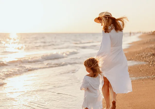 Une mère et son enfant, tous deux en blanc, marchant sur une plage de sable au coucher du soleil, avec les vagues caressant le rivage. Une scène idyllique pour des vacances en famille dans un beach hotel en Guadeloupe.