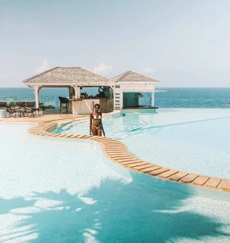 A woman in a yellow swimsuit is standing in an infinity pool with a wooden path, facing a bar and the turquoise sea, representing the ambiance of a 5-star luxury hotel in Guadeloupe.