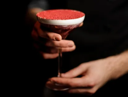 Hands holding an elegant Caribbean cocktail with a white rim and bright red powder on top, suggesting the expertise of a cocktail bar or rum shop in a luxury hotel in Guadeloupe.