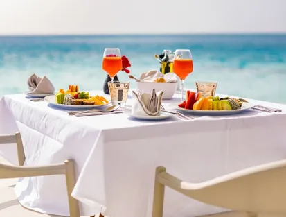 A white table with two seats, fresh fruit dishes and orange cocktails, on a terrace by the sea with the turquoise ocean in the background. This represents a luxury restaurant overlooking the sea in Guadeloupe.