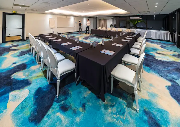 Spacious and modern meeting room arranged in a U-shape, with black tables and white chairs, on a carpet with blue and turquoise patterns, perfect for conferences in a hotel in Guadeloupe.
