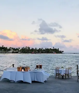 Two tables elegantly set in white on a waterfront pier, with palm trees and a pastel pink and blue sunset in the background, suggesting an idyllic setting for a Caribbean wedding in Guadeloupe or a seaside restaurant.