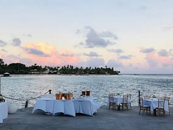 Tables set with white tablecloths and elegant chairs on a waterfront pier at sunset, creating a romantic atmosphere for a Caribbean wedding in Guadeloupe.