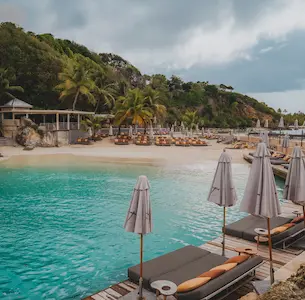 Sandy beach in Guadeloupe with sunbeds and umbrellas on a pier and the sand, turquoise water and green hills with buildings in the background, ideal for a luxury beach hotel in Guadeloupe.