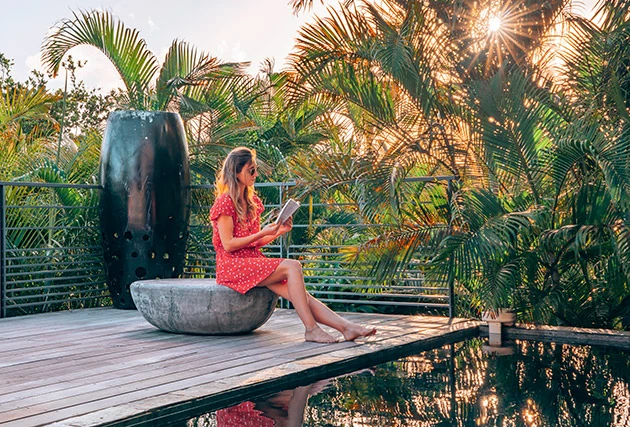 Woman sitting by the pool of a luxury villa with pool in Guadeloupe, surrounded by lush vegetation, reading a book at sunset.