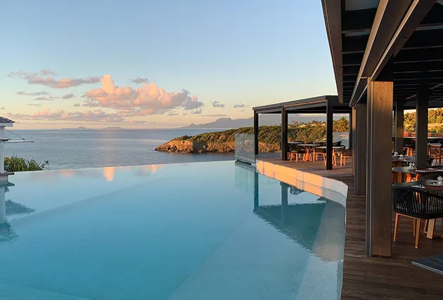 Large infinity pool of a 5-star luxury hotel overlooking the Caribbean Sea at sunset, bordered by a wooden terrace and an outdoor restaurant, located in Sainte-Anne in Guadeloupe.