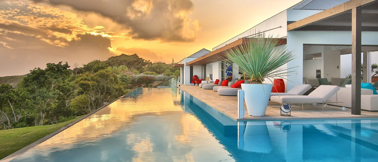 Long infinity pool reflecting an orange and cloudy sunset sky, lined with sun loungers and the facade of a luxury hotel in Guadeloupe, offering breathtaking views of the sea.