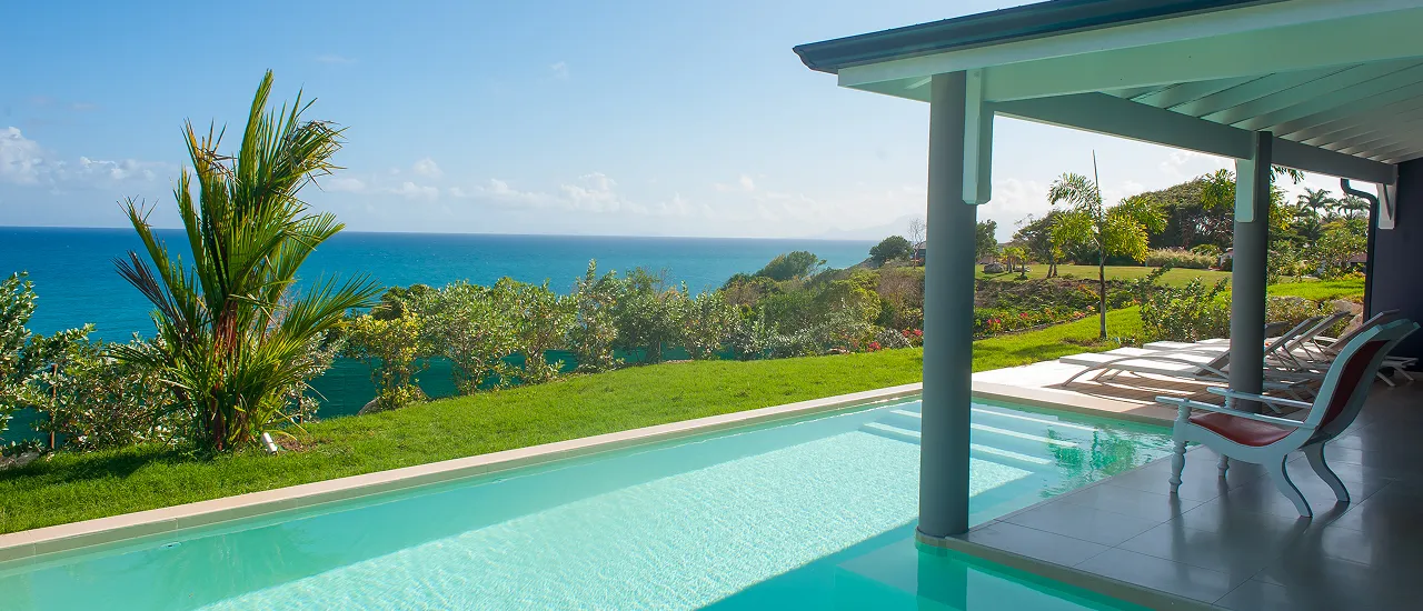 Private pool of a luxury villa overlooking the turquoise sea and a green garden, under a clear blue sky, offering an exclusive experience in Guadeloupe.