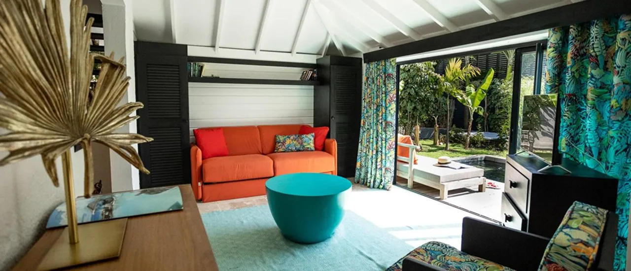 Spacious interior of a luxury bungalow in Guadeloupe, with an orange sofa and a turquoise coffee table, opening onto a private terrace with a small pool and a tropical garden, ideal for renting a villa with pool.