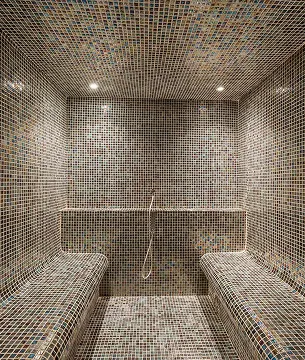 Interior of a luxurious steam room or steam bath, entirely tiled with small mosaics, with two curved benches and a shower, ideal for body treatments and relaxation in a hotel spa in Guadeloupe.