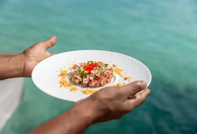 Hands presenting a plate of fresh fish tartare, topped with goldfish eggs, on a turquoise water background. Represents a Creole dish served in a luxury restaurant by the sea in Guadeloupe.