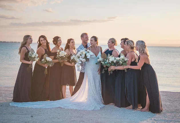 A group of bride and groom and bridesmaids posing on a beach at sunset, the bride in a white dress and the bridesmaids in dark dresses, perfect for a Caribbean wedding in Guadeloupe.
