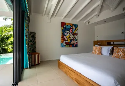 Large, bright bedroom in a luxury villa with a wooden bed, orange patterned cushions, a colorful piece of art on the wall, and large sliding doors opening onto a private pool and vegetation, ideal for renting a villa with pool in Guadeloupe.