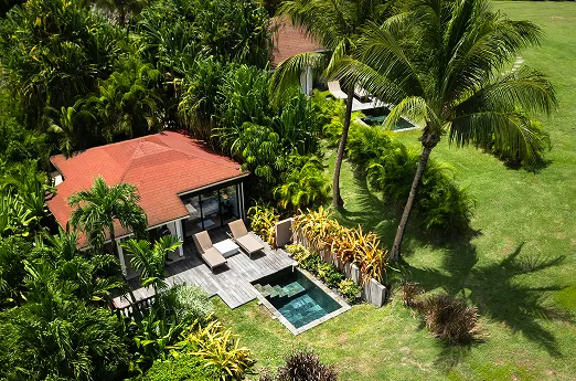 Aerial view of a luxury bungalow with a red roof, a private pool and a wooden deck, surrounded by lush green lawns and palm trees in Guadeloupe, ideal for a villa rental with a pool.