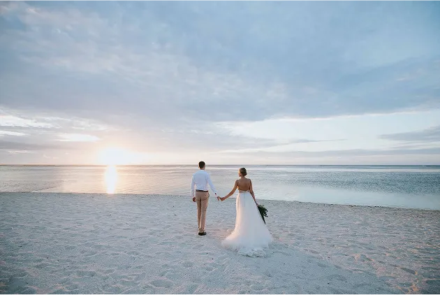 A bridal couple walking hand in hand on a white sand beach at sunset, the bride wearing a long dress. Ideal for a wedding in the Caribbean, in Guadeloupe, in a beach hotel setting.