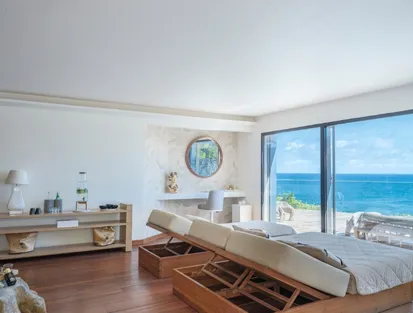 Large bright bedroom in a luxury villa overlooking the sea in Guadeloupe, with a comfortable bed, an open storage bench at the foot of the bed and floor-to-ceiling windows overlooking the turquoise ocean.