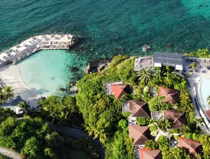 Aerial view of a luxury hotel in Guadeloupe with several bungalows with red roofs, nestled in lush vegetation, overlooking a white sandy beach and a cove with turquoise water, perfect for a 5-star stay in Sainte-Anne.