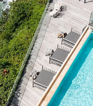 Aerial view of a luxury hotel in Guadeloupe with an infinity pool and a wooden terrace equipped with deckchairs, directly overlooking the turquoise sea and lush coastal vegetation, perfect for a 5-star stay overlooking the sea.