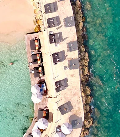 Aerial view of a white sand beach and a cove with turquoise water with a dock equipped with deckchairs and umbrellas, suggesting a luxury 5-star beach hotel overlooking the sea in Sainte-Anne, Guadeloupe.