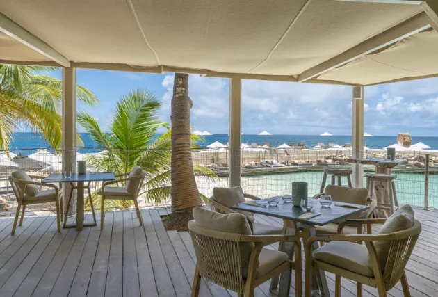 An outdoor bar with a wooden roof and comfortable seating, located near a pool overlooking bungalows and the ocean in the distance. This Caribbean cocktail bar is typical of a luxury hotel in Guadeloupe.