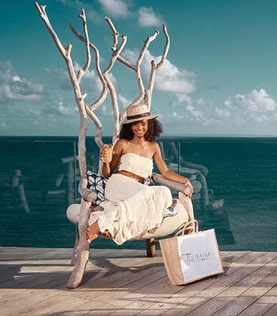 Smiling young woman sitting on a designer wooden chair, holding a cocktail, with a La Toubana Hotel & Spa bag at her feet, on a terrace with panoramic views of the Caribbean Sea