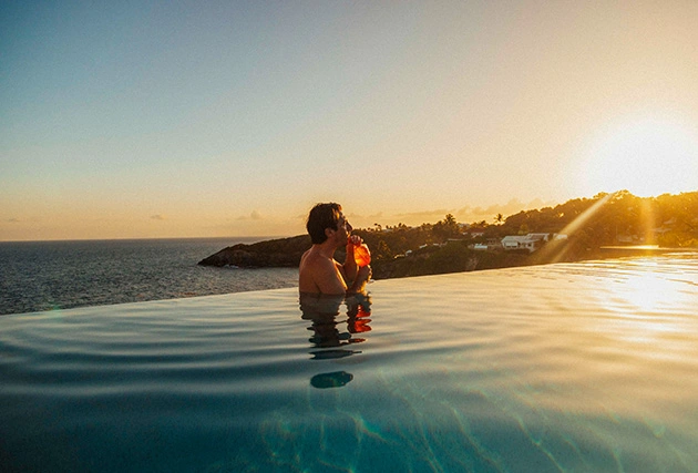 Man enjoying a cocktail in an infinity pool facing the ocean, at sunset in the Islands of Guadeloupe.