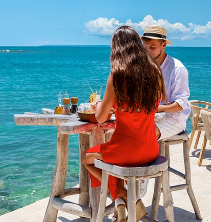 Couple having lunch on the terrace by the sea, at a wooden table facing the turquoise ocean in the islands of Guadeloupe.