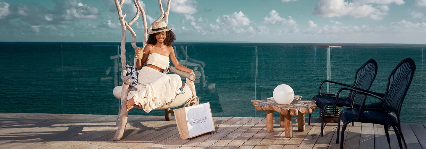 Smiling young woman sitting on a designer wooden chair, holding a cocktail, with a La Toubana Hotel & Spa bag at her feet, on a terrace with panoramic views of the Caribbean Sea